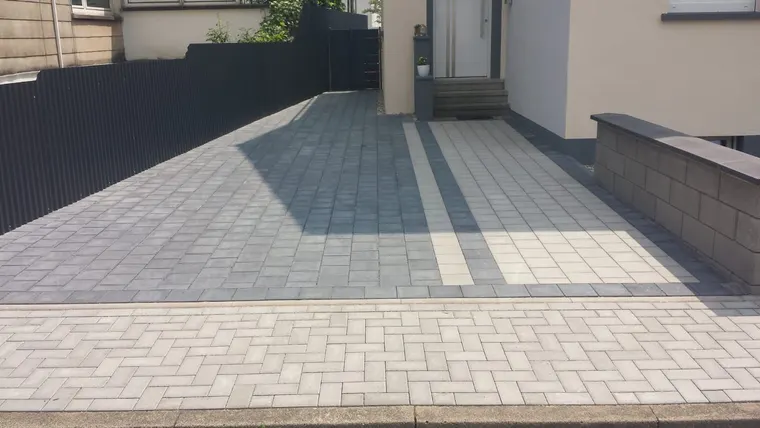 Einfahrt zum Hauseingang aus grauem Pflaster mit hellen Zierstreifen, rechts eine neue Mauer mit Abdeckplatten